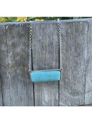 The Linden Turquoise Bar Necklace by Wild Junkie, featuring a rectangular turquoise pendant with a slightly mottled appearance, is displayed hanging on a weathered wooden fence. This silver chain necklace is perfect for western-inspired looks.