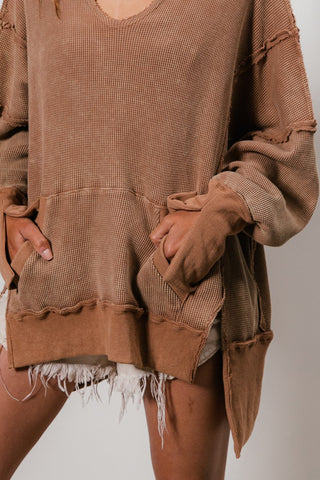 A person is wearing the Blaise Kangaroo Pocket Washed Waffle Thermal Hoodie by BiBi, which features a loose fit, visible seam details, and frayed edges for a distressed look. It has a cozy oversized feel with a large front pocket perfect for resting hands, and is paired with frayed white shorts.