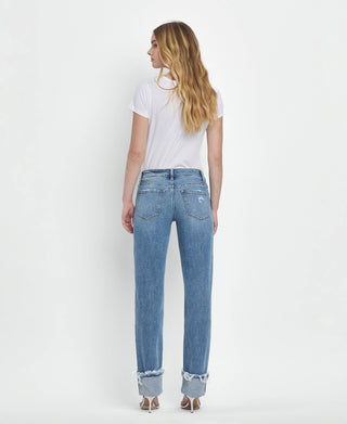 A person with long, wavy blonde hair is standing and facing away from the camera. They are wearing a white t-shirt, Lovervet by VERVET Dezie Mid Rise Cuffed Straight Jeans All Sizes 1-24 in soft stretch denim with distressed hems, and high-heeled sandals. The background is plain white.