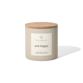 A beige jar labeled "Rich Hippie Candle: 12oz" by Hico Candle Co. features a wooden lid. The hand-poured soy blend candle (12 oz/340 g) emits a captivating fragrance, set against a white background that highlights its minimalist elegance, evoking the subtle bloom of Flower Bomb.