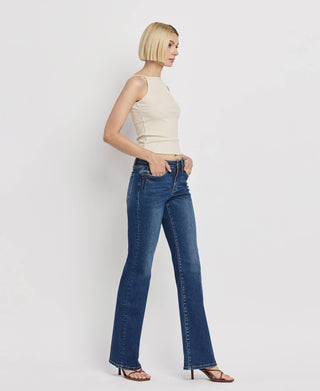 A person with a short blonde bob haircut is posing against a white background. They are wearing a sleeveless beige top, Kip Mid Rise Relaxed Bootcut Jeans by Lovervet from VERVET, and black strappy high-heeled sandals. Their hands are in their pockets as they gaze forward.