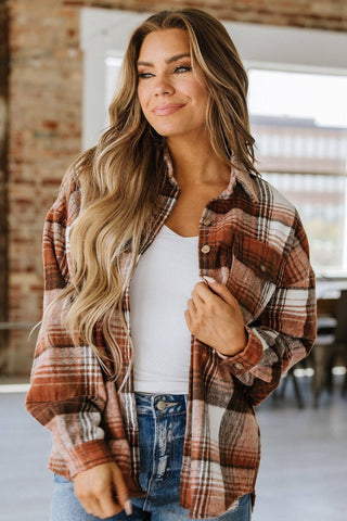 A woman with long hair stylishly wears the Kelsea Plaid Shacket by Liam & Company over a white top and jeans. She stands indoors against a brick wall, gazing thoughtfully to the side.