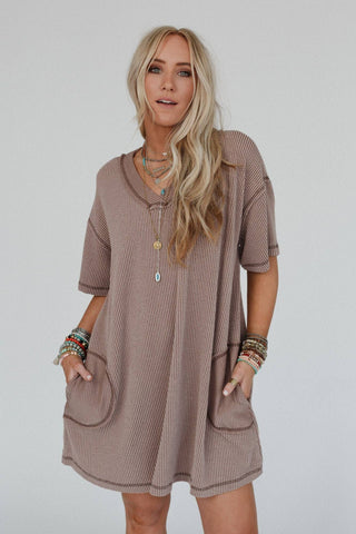 A woman with long, wavy blonde hair is wearing the Brooklyn Soft Touch Tunic Dress by Three Bird Nest. This loose, short-sleeved brown dress features two front pockets. She accessorizes with multiple colorful bracelets, necklaces, and rings while standing against a plain light-colored background.