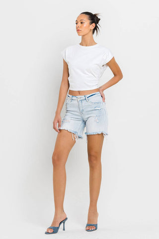 A person stands against a white background, posed with one hand on their hip. They are wearing a white crop top, Kolson Mid Rise Distressed Stretch Denim Shorts by VERVET by FLYING MONKEY, and blue high-heeled shoes. Their hair is tied back in a ponytail.