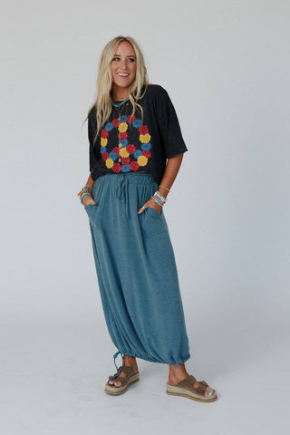 A smiling person with long blonde hair stands against a plain background, wearing a black T-shirt adorned with colorful floral designs, the Rylie Drawstring Maxi Skirt in teal boho style by Three Bird Nest, and brown sandals. Both hands are comfortably resting in their pockets, and they are accessorized with multicolored bracelets.