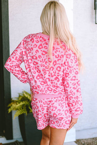 A person with long blonde hair wearing a stylish and comfortable pink and white leopard-print outfit stands with their back to the camera, one hand on their hip. In the background, a potted plant adds natural decor to what appears to be a patio or porch area featuring the Wells 2 Piece Leopard Loungeseat by Lovesoft.