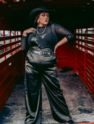 A person stands confidently in a red metal structure, wearing the High Rank Black Mesh Top from Western Edge Boutique, paired with high-waisted pants. The addition of a black cowboy hat, large earrings, and a belt enhances their stylish and bold presence—perfect for a dazzling rodeo night outfit.