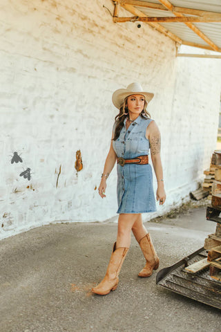 A person in a 2 Fly Denim Sweetheart Dress with Western arrow accents, wearing a cowboy hat and boots, is walking on a concrete path by a rustic white brick wall. Outdoors, they are beside stacked wooden pallets under a corrugated metal roof.