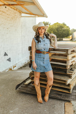 A woman wearing a 2 Fly Denim Sweetheart Dress, cowboy hat, and western boots stands with one hand on her hat next to a stack of wooden pallets outside a building with a white brick wall adorned with two decorative flies.