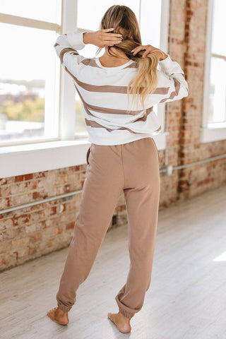 In a bright room with large windows and brick walls, someone stands barefoot in the comfortable Callahan Striped Pullover and Joggers Set by Liam & Company. They're adjusting their long hair with both hands, exuding comfort in their cozy attire.