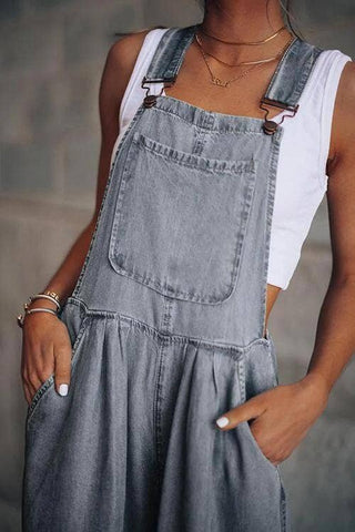 A person wearing a sleeveless white shirt paired with Honeypeach's Everson Lightweight Denim Wide Leg Denim Overall, featuring a front pocket and adjustable shoulder straps. Their hands are casually placed in the overall's side pockets, accessorized with a gold necklace and bracelet.