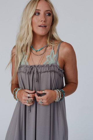 A blonde woman stands against a plain background wearing the Three Bird Nest's Heavenly Glow Maxi Dress in Mushroom, adorned with delicate lace detailing, exuding romantic charm. She accessorizes with layered turquoise necklaces, bracelets, and rings on both hands. Her hair is styled in loose, natural waves as she gazes to the side.