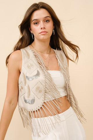 A person with long hair wears the Faux Leather Embellished Fringe Vest from Western Edge Boutique over a white tube top and high-waisted pants. The vest features decorative patterns that accentuate the western style as the individual stands against a plain, light-colored background.