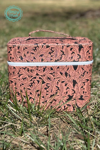 An essential Heard your Makeup Tool Box from Western Edge Boutique with a square, floral-patterned design in pink and black rests on the grass. This exquisite box boasts a convenient top handle and a white zipper encircling its center, making it perfect for stylish organization.