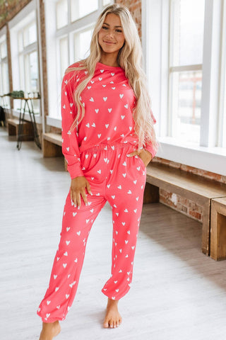 A person with long blonde hair is smiling while wearing the Lasso My Heart Red White Hearts Print Lounge Set by Liam & Company. They stand barefoot in a bright room with large windows.