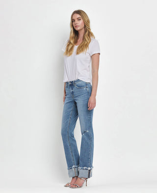 A person with long blonde hair stands against a white background. They are wearing a white T-shirt, Lovervet by VERVET's Dezie Mid Rise Cuffed Straight Jeans All Sizes 1-24 with frayed hems, and silver high-heeled sandals. The person is standing with one hand by their side and the other slightly bent.