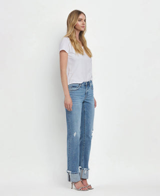 A woman with long blonde hair is standing and facing to the side, wearing a plain white t-shirt, Lovervet by VERVET Dezie Mid Rise Cuffed Straight Jeans in light blue soft stretch denim with distressed flair at the ankles, and high-heeled sandals. The background is plain white.