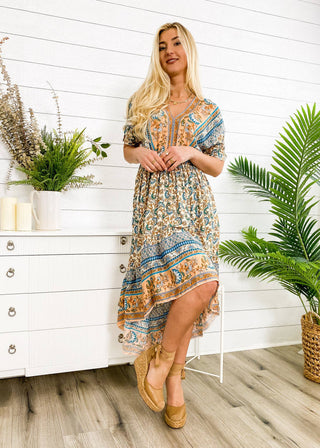 A woman with long blonde hair stands indoors, wearing the Finn Tie Back High Low Dress from Anna Kaci Wholesale. The dress features a flowing, floral print in blue, orange, and cream. She has one leg slightly raised as she leans against a white dresser adorned with a plant. The setting includes green potted plants and light-toned wooden flooring.