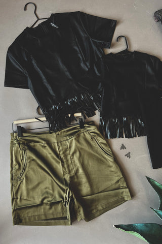 A flat lay featuring two black tops with fringe details at the bottom, along with a pair of 2 Fly's Lawsen Tuxedo Shorts in olive green, complete with a drawstring and functional pockets. The larger top is displayed on a hanger with a brand tag attached. These items are set against a plain background, accented by two small butterfly decals nearby.