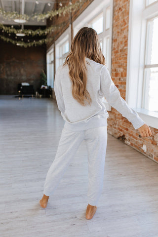 A person with long hair, wearing a cozy two-piece Alison Half Zip Loungewear Set from Liam & Company, stands barefoot in a spacious room with light wooden floors, exposed brick walls, large windows, and hanging greenery. They are facing away from the camera.