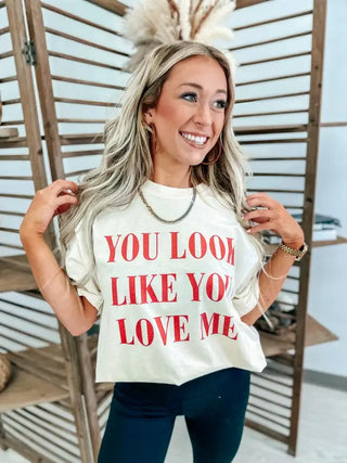 A woman with long hair smiles, holding her hair back with both hands. She's wearing Bird & Co Wholesale Tees' "You Look Like You Love Me" ivory graphic tee. The Comfort Colors shirt sets a relaxed tone as she stands in front of wooden shelves.