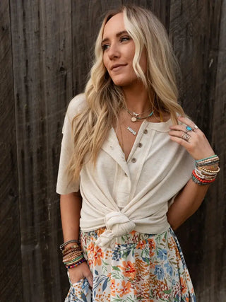 A woman with long blonde hair stands against a wooden wall, wearing the Soft Plume Henley Top from Western Edge Boutique paired with a colorful floral skirt. She accessorizes with several necklaces and bracelets, gazing off to the side with a relaxed expression, exuding an effortless charm in her comfortable fabric ensemble.