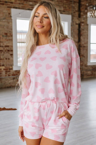 A person with long blonde hair is indoors in a brick-walled room with large windows, wearing the Sammy Heart Print Loungewear Set by Liam & Company. They casually tuck their hands into the shorts' pockets.