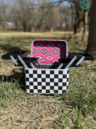 A "Heard your Makeup Tool Box" by Western Edge Boutique, showcasing its checkered exterior and pink interior with a geometric pattern, is open on the grass in a wooded area. Sunlight casts shadows on the ground, accentuating the must-have case's extendable trays.