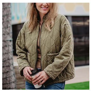 A person wearing a Kipton Quilted Jacket in olive green from Western Edge Boutique and blue jeans holds a disposable coffee cup with both hands. They are outside, standing near a tree, with a building and mural in the background. The person's face is partially visible.