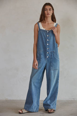 A woman with long brown hair stands against a plain background, donning the Kodi Denim Square Neck Button-down Wide Leg Overalls: Denim by By Together, layered over a button-down sleeveless white tank top. The overalls feature large front pockets and button closures down the front. She completes her look with open-toed flat sandals.