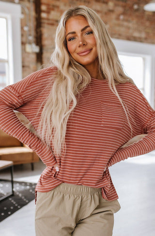 A smiling woman with long blonde hair looks at the camera, wearing a tailored-fit red and white Liza Striped Long Sleeve Top from Liam & Company, paired with beige pants. The background reveals a casually furnished room featuring brick walls and large windows.