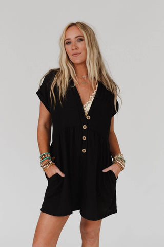 A woman with long blonde hair is wearing the Sage and Wildflowers Romper in black by Three Bird Nest, which is made of comfortable knit fabric. She has her hands in the romper's pockets and is accessorized with multiple stacked bracelets on both wrists, embodying a boho-chic vibe against a plain white background.