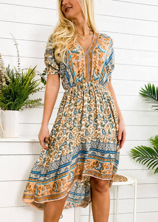 A woman with long blonde hair is wearing the Finn Tie Back High Low Dress from Anna Kaci Wholesale, featuring a knee-length floral print in a blend of blue, orange, and beige hues. She stands next to a white cabinet adorned with potted plants, set against a backdrop of a white paneled wall.