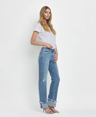 A person posing against a plain white background is wearing a white t-shirt tucked into Lovervet by VERVET's Dezie Mid Rise Cuffed Straight Jeans All Sizes 1-24, made of soft stretch denim. They are also sporting silver high-heeled sandals. With long, wavy hair, they stand in a relaxed stance with one hand on their hip.