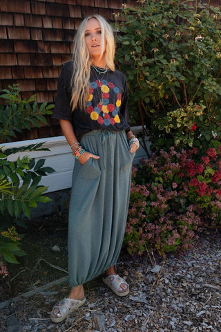 Standing in front of a house with wooden siding and a lush garden, a woman with long blonde hair wears a black t-shirt adorned with colorful circular patterns. She pairs it with the Rylie Drawstring Maxi Skirt by Three Bird Nest in teal, which features an elastic drawstring waistline. Her look is accessorized with multiple bracelets and necklaces, and she finishes it off with sandals.