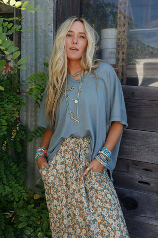 A woman with long blonde hair stands outdoors in front of a rustic wooden door and lush greenery. Embracing a bohemian style, she wears the Payton Top from Three Bird Nest, a loose gray shirt made of stretchy knit fabric, paired with a long floral skirt. She accessorizes with multiple layered necklaces and blue stacked bracelets. Her hands rest in her skirt pockets as she looks at the camera.