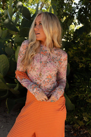 A woman with long blonde hair stands in front of a background of large green cactus plants. She is wearing the Made You Blush Floral Top - Pink Floral by Three Bird Nest, paired with a high-waisted, orange skirt. She gazes off to the side, one hand resting near her face and the other on her skirt.