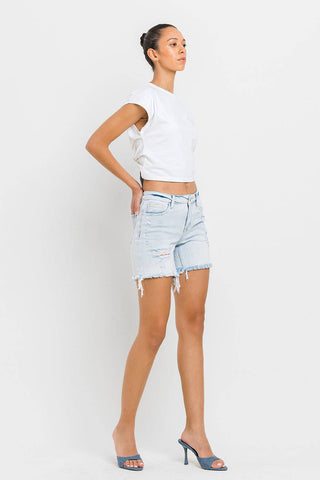 A person stands against a plain white background, wearing a white sleeveless top, the Kolson Mid Rise Distressed Stretch Denim Shorts by VERVET by FLYING MONKEY in light blue, and blue high-heeled sandals. Their hair is pulled back neatly, and they are posed in a profile view with one hand on their hip.