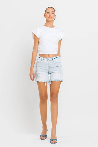 A person with hair tied back stands against a plain white background, wearing a white t-shirt and VERVET by FLYING MONKEY's Kolson Mid Rise Distressed Stretch Denim Shorts in light blue. They are also sporting blue open-toe heels. The individual's expression is neutral.