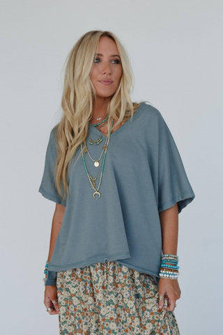 A blonde woman with long hair wears the Payton Top – a loose, blue-gray stretchy knit piece by Three Bird Nest. She accessorizes her look with multiple layered necklaces and a flowy, floral skirt that perfectly embodies bohemian style. Several bracelets adorn both of her wrists as she stands against a plain, light background, smiling slightly.