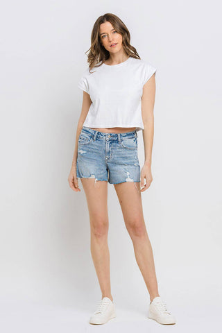 A woman stands against a plain white background. She wears a white, short-sleeved crop top and the VERVET by FLYING MONKEY Kylie Mid Rise Raw Distressed Hem shorts, available in both regular and curvy sizes. Her hair is shoulder-length and wavy, and she has a relaxed expression with her arms by her sides.