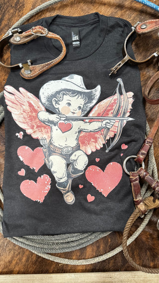 Western Edge Boutique's "Cowboy Cupid" tee features a cherub in a cowboy hat and boots, aiming its bow and arrow. This Valentine's Day design includes wings flanked by pink hearts, coiled ropes, and leather straps for a rustic touch.