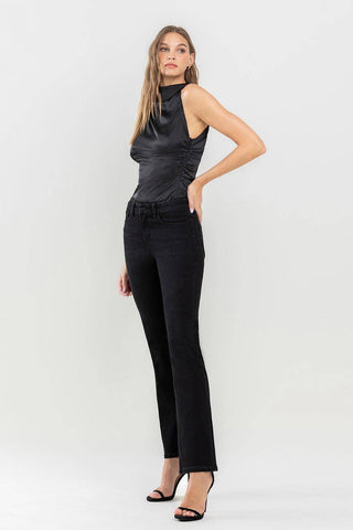 A woman stands against a plain background, wearing a sleeveless black top with ruching on the sides and Halston Mid Rise Bootcut Jeans from Lovervet by VERVET. She has long hair and is posing with one hand on her hip. She completes her wardrobe must-have look with black heeled sandals.