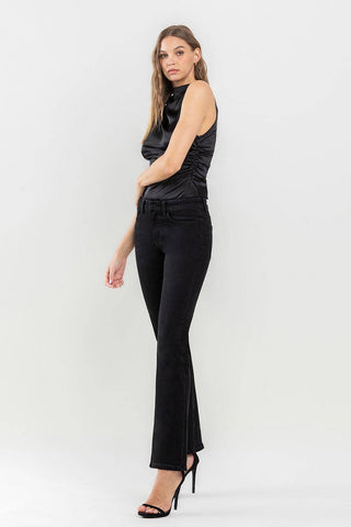 A woman with long hair wears a sleeveless black top and the Halston Mid Rise Bootcut Jeans by Lovervet by VERVET, a wardrobe must-have. She stands in a relaxed pose with one hand touching her arm and one leg slightly forward. She wears black high-heeled sandals against a plain white background.