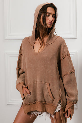 A woman stands against a white paneled wall, wearing the Blaise Kangaroo Pocket Washed Waffle Thermal Hoodie by BiBi, featuring distressed edges and a kangaroo pocket. Her long hair is loose, and she gazes slightly to the side. She completes the casual look with frayed shorts.