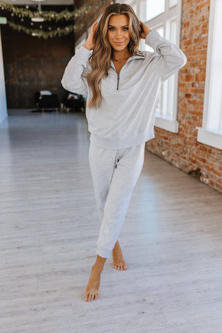 A woman stands barefoot in a bright room with brick walls and large windows. She is wearing a cozy Liam & Company Alison Half Zip Loungewear Set in light gray, featuring a long-sleeve zip-up top and matching jogger pants. With long wavy hair, she touches the sides of her head while smiling at the camera.