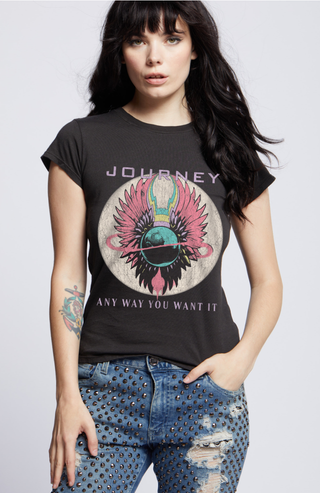 A woman with long black hair stands against a plain background, showcasing her rocker-inspired look. She is wearing a vintage black "Journey Any Way You Want It Only the Young Baby Graphic Tee" from Recycled Karma, made of 100% soft cotton. Her ripped jeans feature a studded design, and she has a visible tattoo on her left forearm.