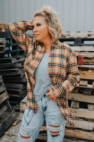 A person with short, curly blonde hair stands in front of a stack of wooden pallets. They are sporting a One24 Harvest Flannel plaid shirt over a gray t-shirt and distressed jeans with multiple rips. One hand is resting on their head, and the other is tucked into their pocket, embodying the perfect fall makeover for the autumn season.