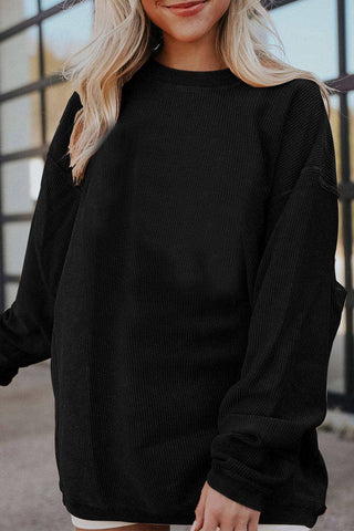 A person with long, blonde hair wearing a Liam & Company Asia Ribbed Oversized Sweatshirt stands in front of a building with large glass windows. They are smiling and have their left hand slightly behind them, showcasing the plush ribbed fabric of their cozy attire.