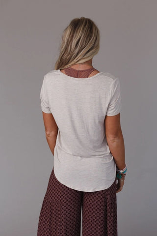 A woman stands facing away from the camera, wearing a white Moxen Scoop Neck Bralette Tee from Three Bird Nest with a curved hemline and patterned maroon pants. She has long, wavy blonde hair and is accessorized with multiple bracelets on her left wrist. The background is plain and gray.